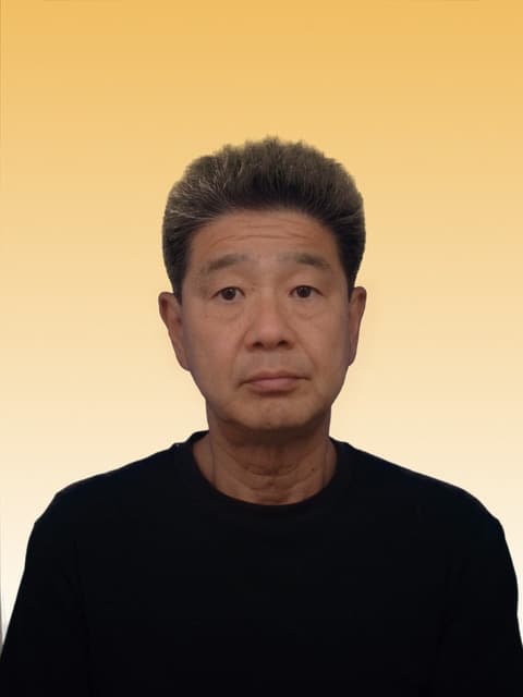 代表・新飯田啓二の顔写真（黒のトップス、明るいベージュ調の背景）